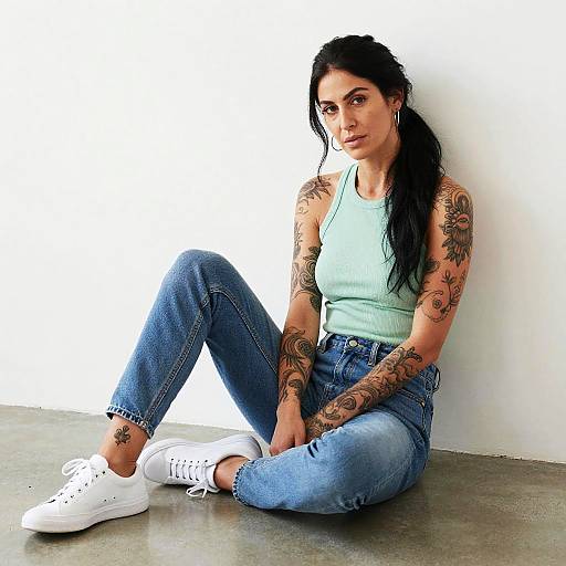 Casual Woman with Tattoos Sitting Against White Wall in Light Green Top and Blue Jeans