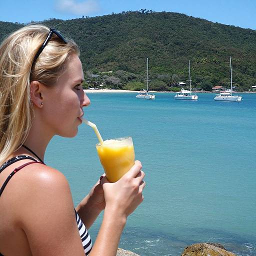 Blonde woman in striped bikini sips orange drink by turquoise water, sailboats, and green hills in background; bright sunny day.