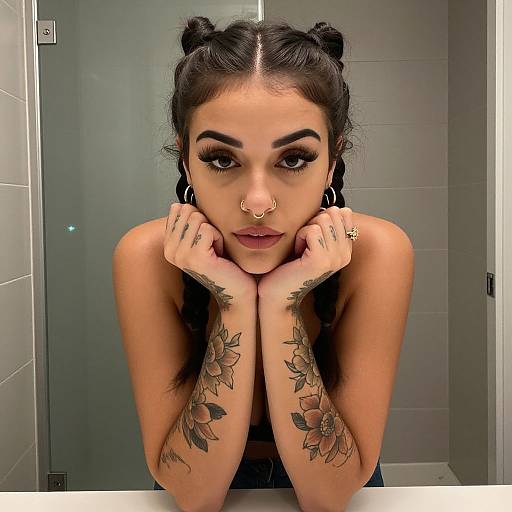 Photograph of a tattooed woman with dark hair in buns, large eyelashes, nose ring, floral tattoos on arms, hands resting on chin