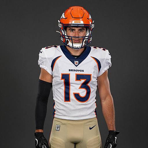 Photograph of a muscular male football player in white Denver Broncos jersey (#13), orange helmet, beige pants, black gloves, and black arm sleeve,