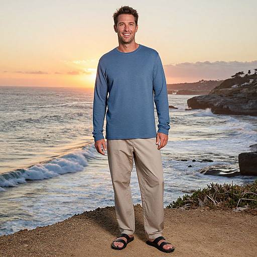 Photograph of a smiling man with short brown hair, blue long-sleeve shirt, beige pants, black sandals, standing on a rocky beach at