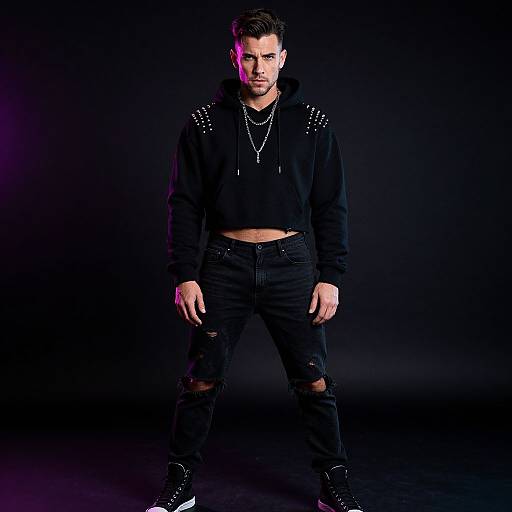 Photograph of a young, attractive man with short brown hair, wearing a black studded hoodie, ripped jeans, and black shoes, standing against a