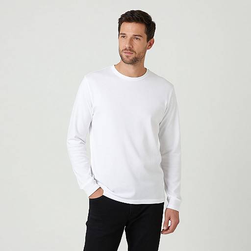 Photograph of a bearded man with short dark hair, wearing a plain white long-sleeve shirt and black pants, standing against a plain white