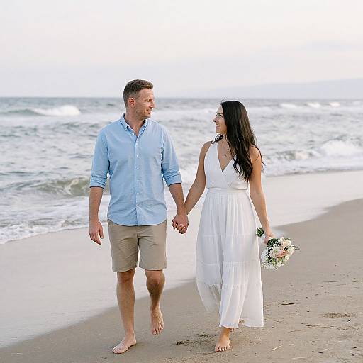 Photograph of a smiling couple holding hands on a beach; he in a light blue shirt and beige shorts, she in a white dress, carrying flowers