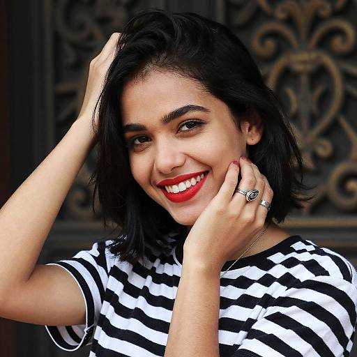 Photograph of a smiling woman with medium skin tone, black shoulder-length hair, wearing a black-and-white striped shirt, red lipstick, silver ring,