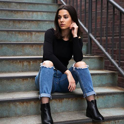 Photograph of a young woman with dark hair, black long-sleeve top, ripped blue jeans, fishnet stockings, and black boots, sitting