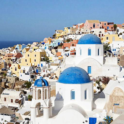 Photograph of a vibrant, sunlit Greek island village with white buildings and blue domes, set against a clear blue sky and distant sea.