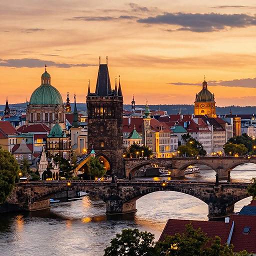 Photograph of Prague at sunset, featuring the Charles Bridge with arches, historic buildings with red roofs, green domes, and a colorful sky.