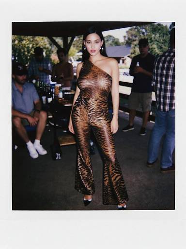 Photograph of a woman with dark hair wearing a one-shoulder, tiger-print jumpsuit, standing outdoors at a casual event with people in the