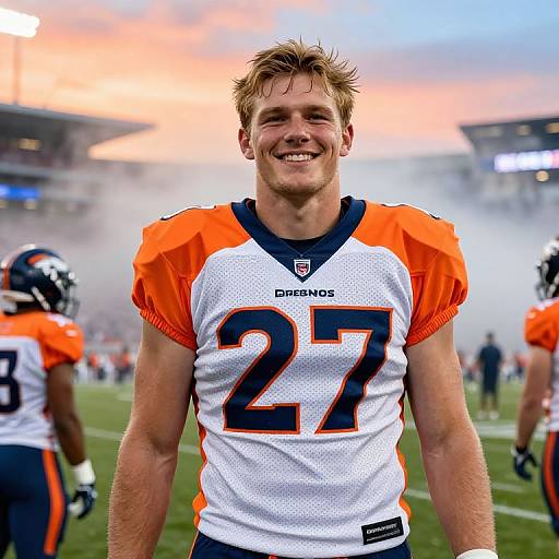 Photograph of smiling NFL quarterback in Denver Broncos jersey number 27, orange and white, on football field at sunset, with teammates and fog in background
