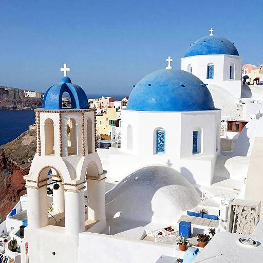 Photograph of a bright, sunlit Greek island with white-washed buildings and blue domes, featuring a small church with cross-topped bell towers