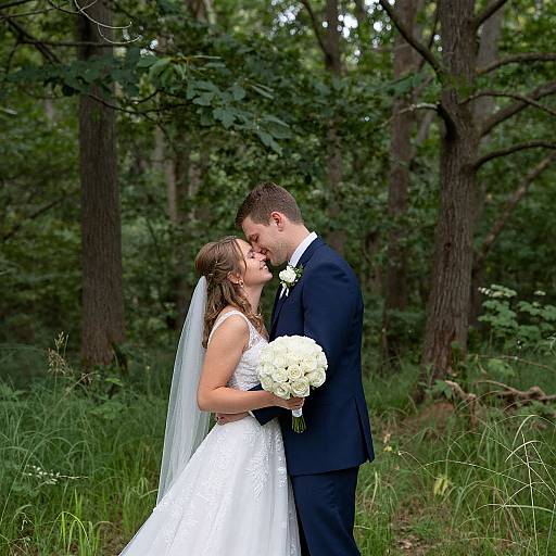 Photograph of a bride in a white lace dress and veil, and groom in a black suit, kissing in a lush, green forest, with the