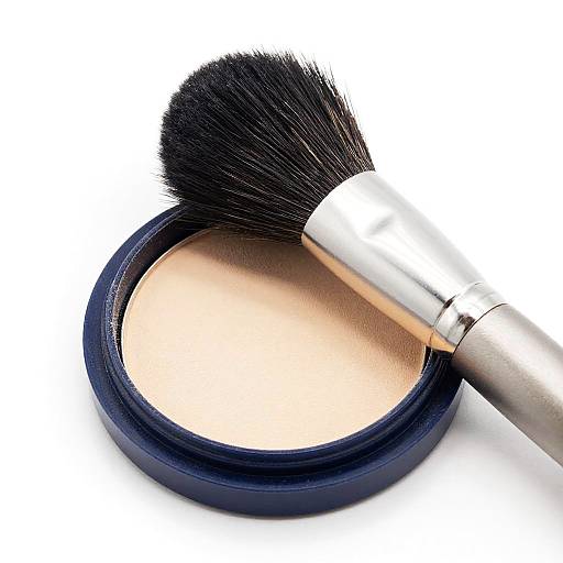 Photograph of a round, black-capped compact with beige powder, partially open, and a metallic brush with black bristles gently touching the powder.