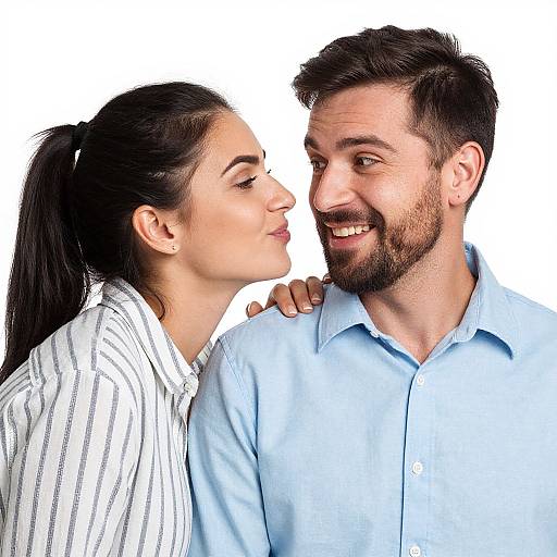 Photograph of a smiling bearded man in a light blue shirt, with a woman in a striped shirt leaning in to kiss his cheek. White background