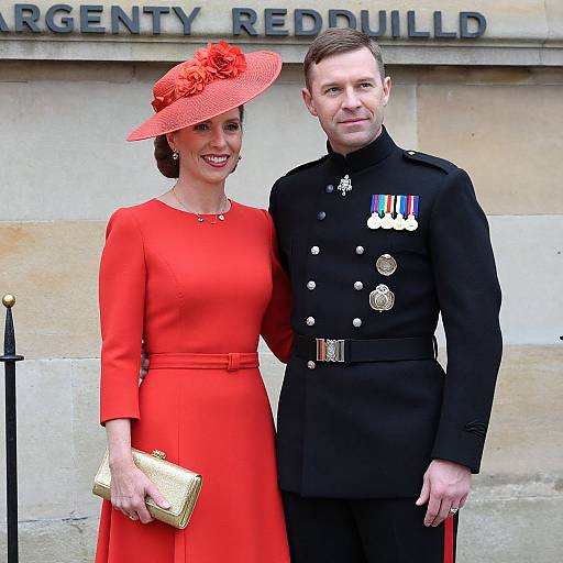 Photograph of a smiling woman in a red dress and hat, holding a gold clutch, standing beside a man in a black military uniform with medals,