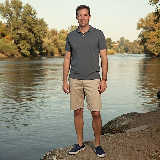 Photograph of a smiling, short-haired man in a dark polo shirt, beige shorts, and navy sneakers standing by a calm river with autumn trees in