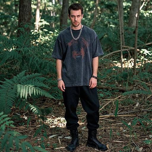 Photograph of a man with short dark hair, wearing a stained gray T-shirt, black pants, and black boots, standing in a forest with fern