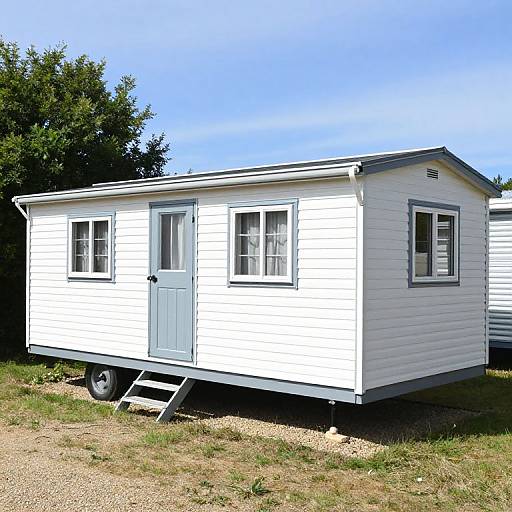 Photograph of a small, white, rectangular trailer with blue trim, two windows, and a door, set on a gravel patch with a ladder,