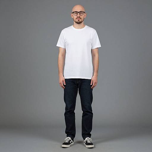 Photograph of a bald, bearded man with glasses, wearing a white t-shirt, black pants, and black sneakers, standing against a gray backdrop