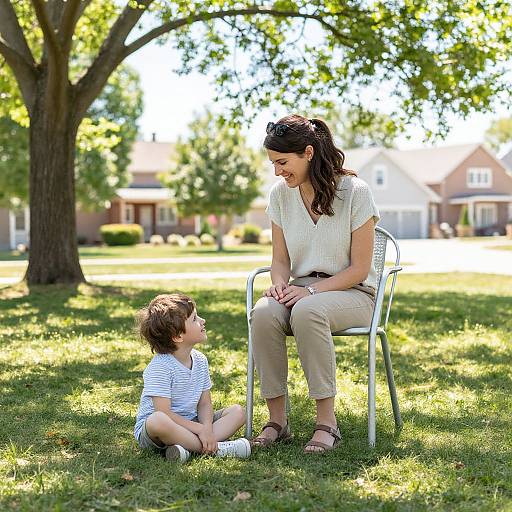 Photograph of a smiling woman with dark hair in a white blouse and beige pants, sitting on a chair, looking at a young boy in a striped