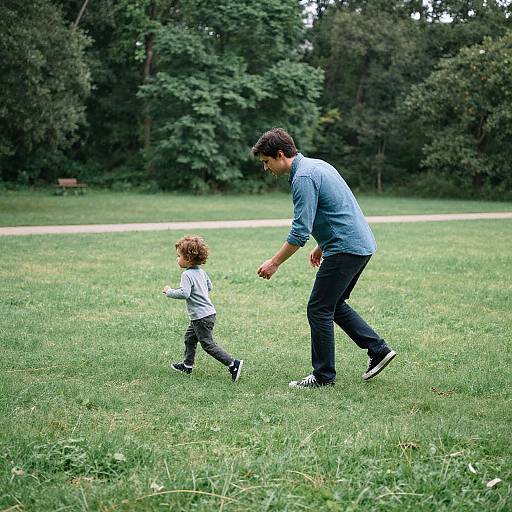 Photograph of a father in a blue shirt and black jeans running on a green grassy field with his curly-haired toddler, both smiling, surrounded by