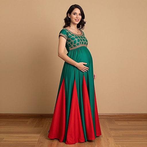 Pregnant woman with curly black hair, wearing a green and red traditional Indian gown with gold embroidery, standing on wooden floor against beige wall. Photograph