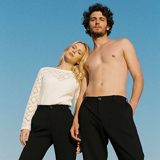 Photograph of a topless, curly-haired bearded man in black pants and a blonde woman with wavy hair in a white lace top, standing
