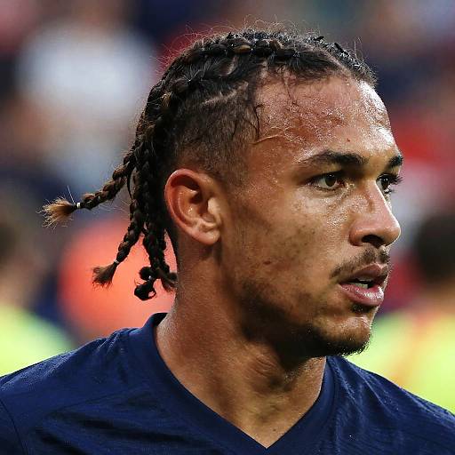 Photograph of muscular, dark-skinned male soccer player with braided hair, black V-neck shirt, focused gaze, and slightly sweaty face in a