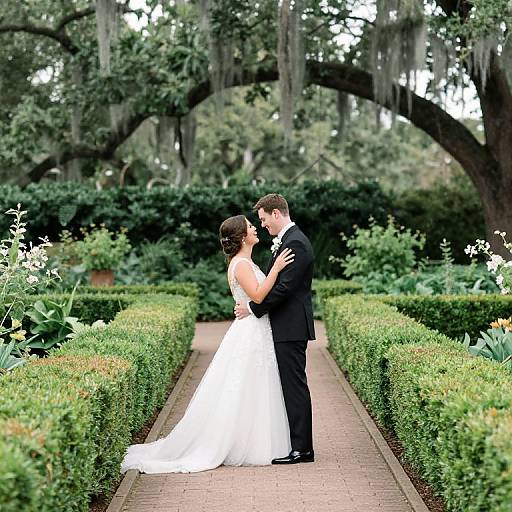 Photograph of a bride in a white gown and groom in a black suit kissing in a lush, green garden with manicured hedges and large trees