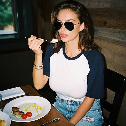 Photograph of a woman with brown hair, wearing black-and-white raglan shirt, ripped jeans, and dark sunglasses, eating with a fork at a