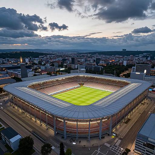 Aerial photograph of a modern football stadium with illuminated green field, surrounded by cityscape, under dramatic cloudy evening sky.