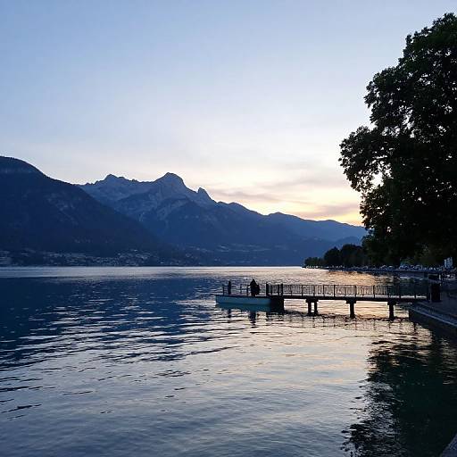 Photograph of a serene lake at sunset, featuring a silhouetted pier, calm water, and mountainous background under a gradient blue-to-white