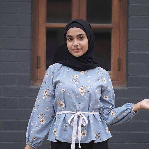 Photograph of a young South Asian woman with medium skin, wearing a black hijab, light blue floral blouse, and white ribbon belt, standing in