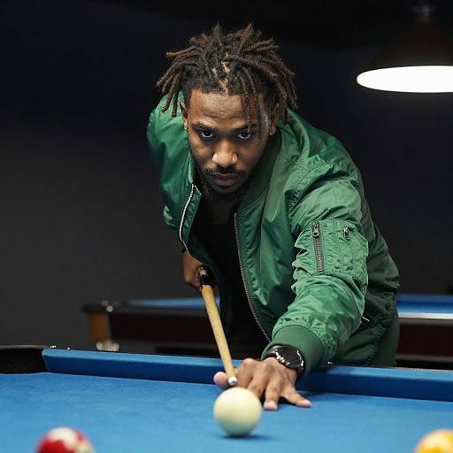 Photograph of a focused Black man with dreadlocks, wearing a green jacket, aiming to strike a billiard ball on a blue pool table.