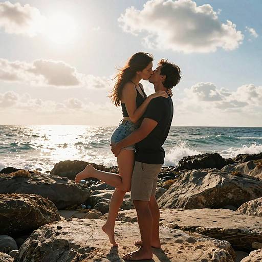 Photograph of a couple kissing on rocky beach at sunset, silhouetted against the sun and ocean waves, with the woman lifted.