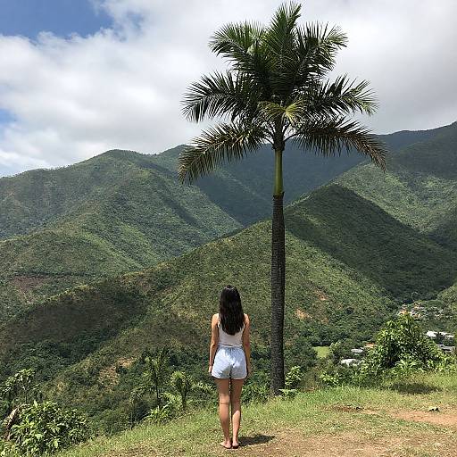 Photograph of a woman with long black hair in a white sleeveless dress, standing barefoot on grass, facing lush green mountains and a tall palm