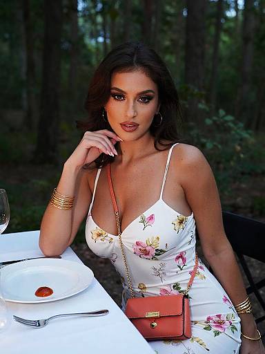 Photograph of a beautiful woman with dark hair, wearing a floral white dress, gold bracelets, and a red purse, seated at an outdoor table in