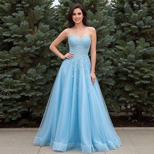 Photograph of a smiling woman with long dark hair in a strapless, light blue, lace-embellished ball gown, standing in front of