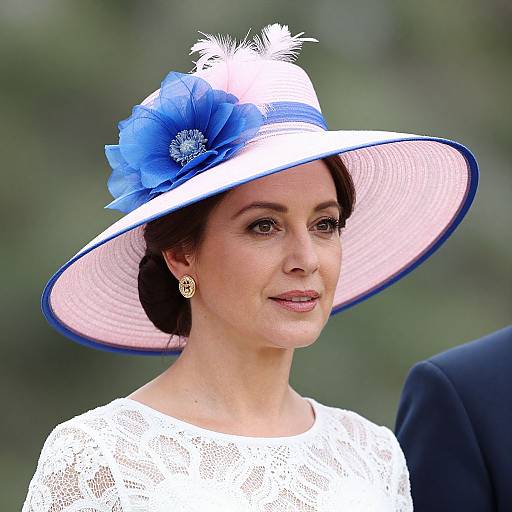 Photograph of a woman with fair skin, dark hair, wearing a white lace top, blue flowered hat with white feathers, and gold earrings,