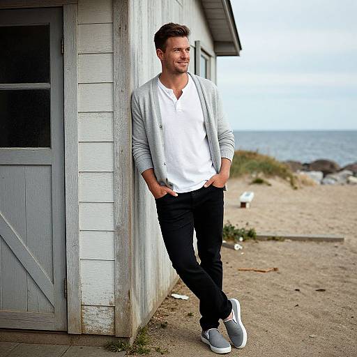 Photograph of a handsome man with short brown hair, smiling, leaning against a weathered white beachside shed in casual white shirt, gray cardigan
