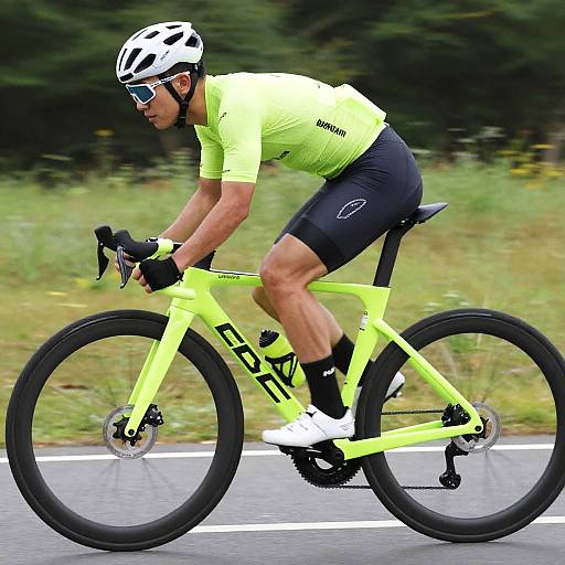 Photograph of a male cyclist in a neon yellow and black outfit, white helmet, and sunglasses, riding a bright yellow bike on a road. Green