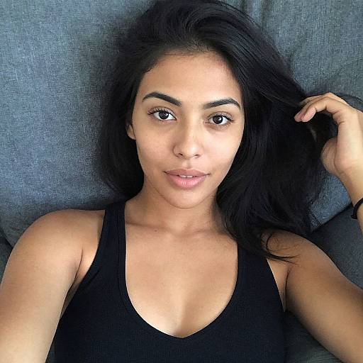 Photograph of a young woman with long black hair, brown eyes, and olive skin, wearing a black tank top, lying on a blue-gray couch