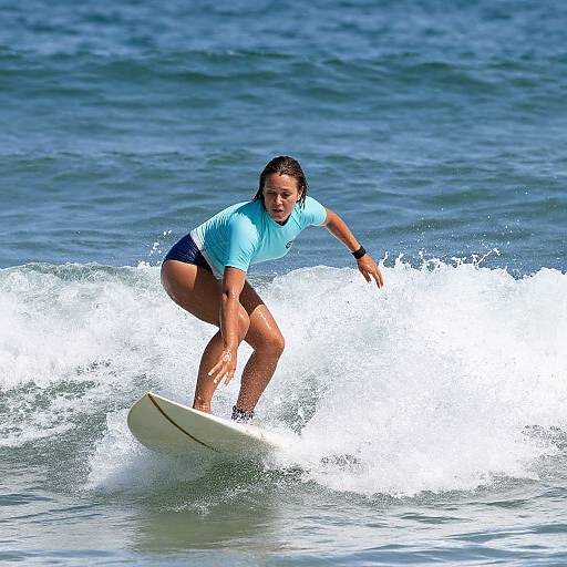 Photograph of a tan-skinned woman with wet brown hair, wearing a light blue shirt and black shorts, surfing on a small wave in the blue