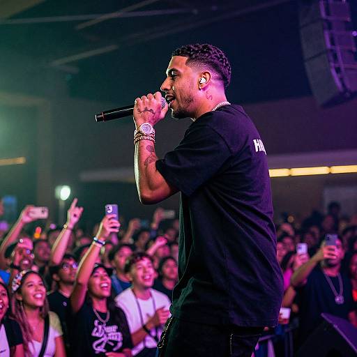 Photograph of a male rapper with tattoos, black t-shirt, and wristbands, singing into a microphone on stage, crowd cheering, holding phones,