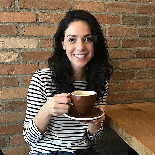 Photograph of a smiling woman with long black hair, wearing a black-and-white striped shirt, holding a brown coffee cup, seated at a wooden table