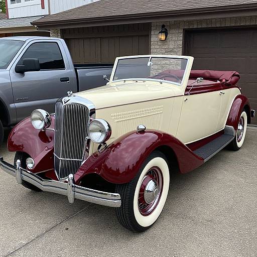 Photograph of a classic maroon and cream vintage convertible with white-walled tires, parked in a driveway next to a gray truck.