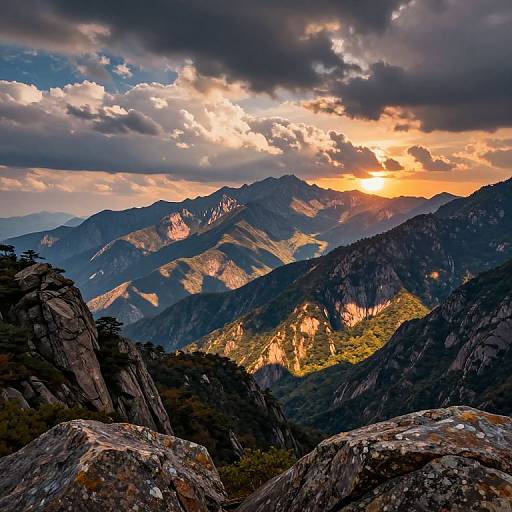 Photograph of a dramatic mountain range at sunset, with vibrant orange sunlight illuminating the peaks, dark clouds overhead, and rocky foreground.