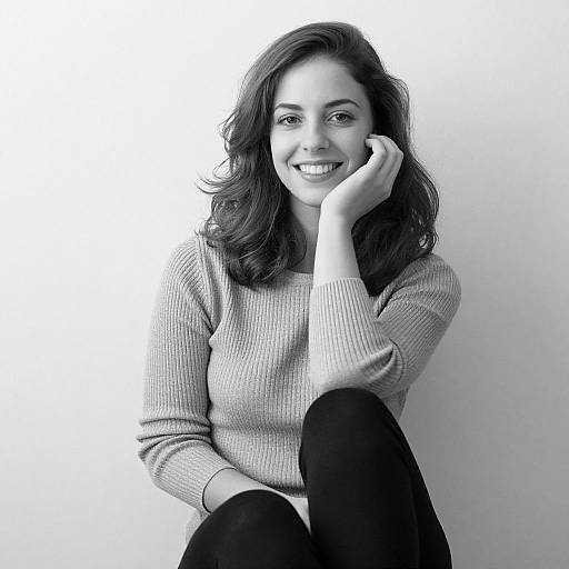 Black-and-white photograph of a smiling woman with wavy dark hair, wearing a ribbed sweater and black pants, resting her chin on her hand against
