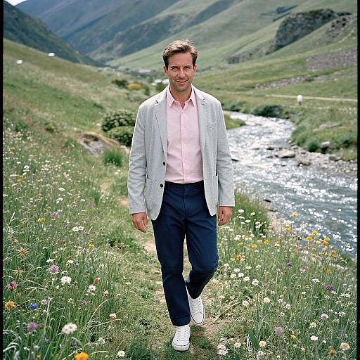 Photograph of a smiling man in a pink shirt, light jacket, and dark pants, walking through a sunny, flower-filled meadow by a shimmer