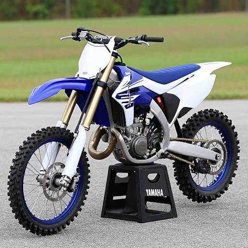 Photograph of a blue and white motocross bike with black tires, gold fork, and exhaust, standing on a black Yamaha block.